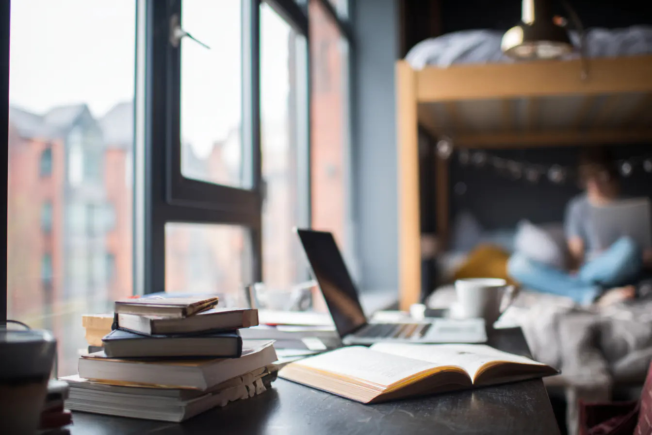 Stapel Bücher, Laptop und offenes Buch auf einem Tisch vor einem Fenster. Im Hintergrund eine Person auf einem Bett.